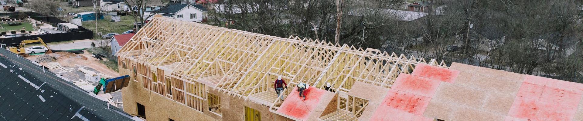Aerial view of a long timber-framed building with roof sheathing going on